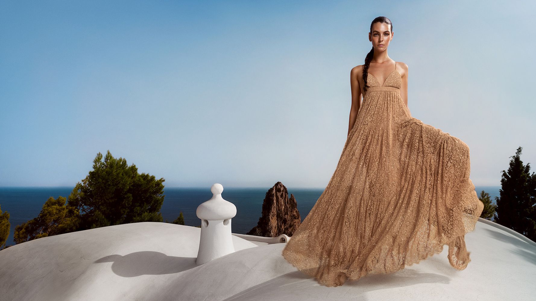 A model in a flowing golden lace dress on a whitewashed terrace above the sea